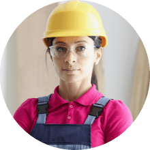 Serious female construction worker wearing a pink shirt, overalls, yellow hard hat, and safety glasses.