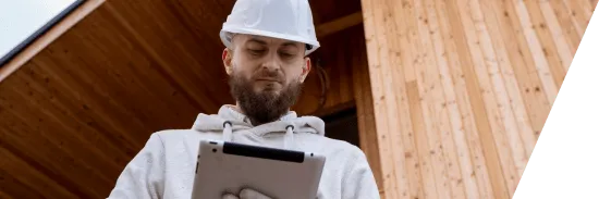 Bearded construction worker in a white hard hat using a tablet in front of a wooden building.