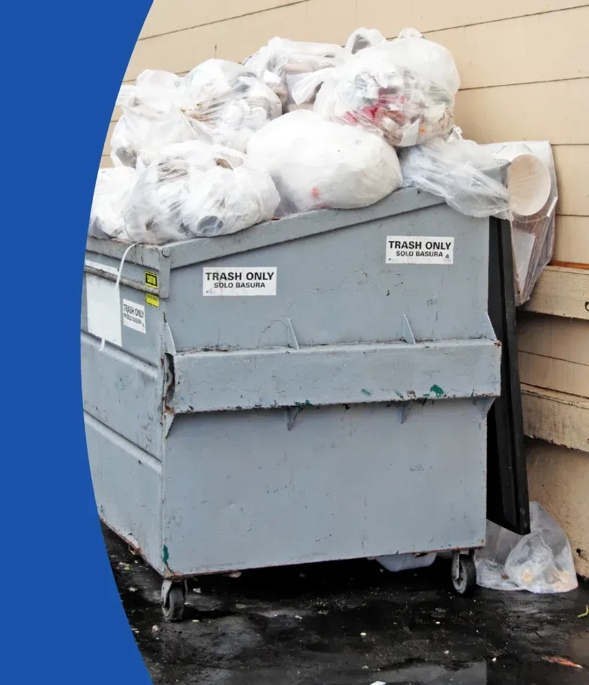 A large gray dumpster with "Trash Only" labels on its sides, overflowing with plastic garbage bags while sitting on a wet surface.