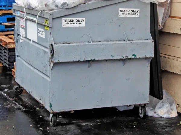 A large gray dumpster with "Trash Only" labels on its sides, overflowing with plastic garbage bags while sitting on a wet surface.