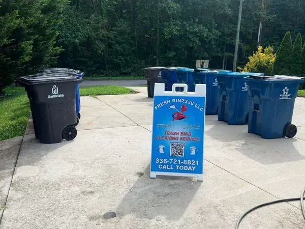 A "Fresh Binz 336 LLC" A-frame sign advertising trash bin cleaning service, surrounded by multiple blue and black bins.