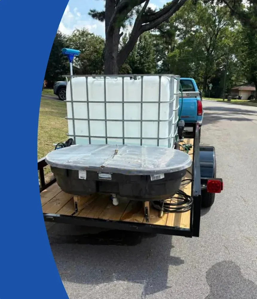 A utility trailer carrying a large caged water tank and a smaller flat tank, parked on asphalt near a blue truck.