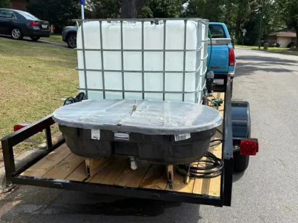 A utility trailer carrying a large caged water tank and a smaller flat tank, parked on asphalt near a blue truck.