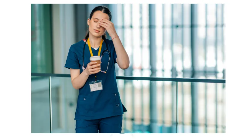 Tired healthcare worker in blue scrubs holding coffee and rubbing her forehead