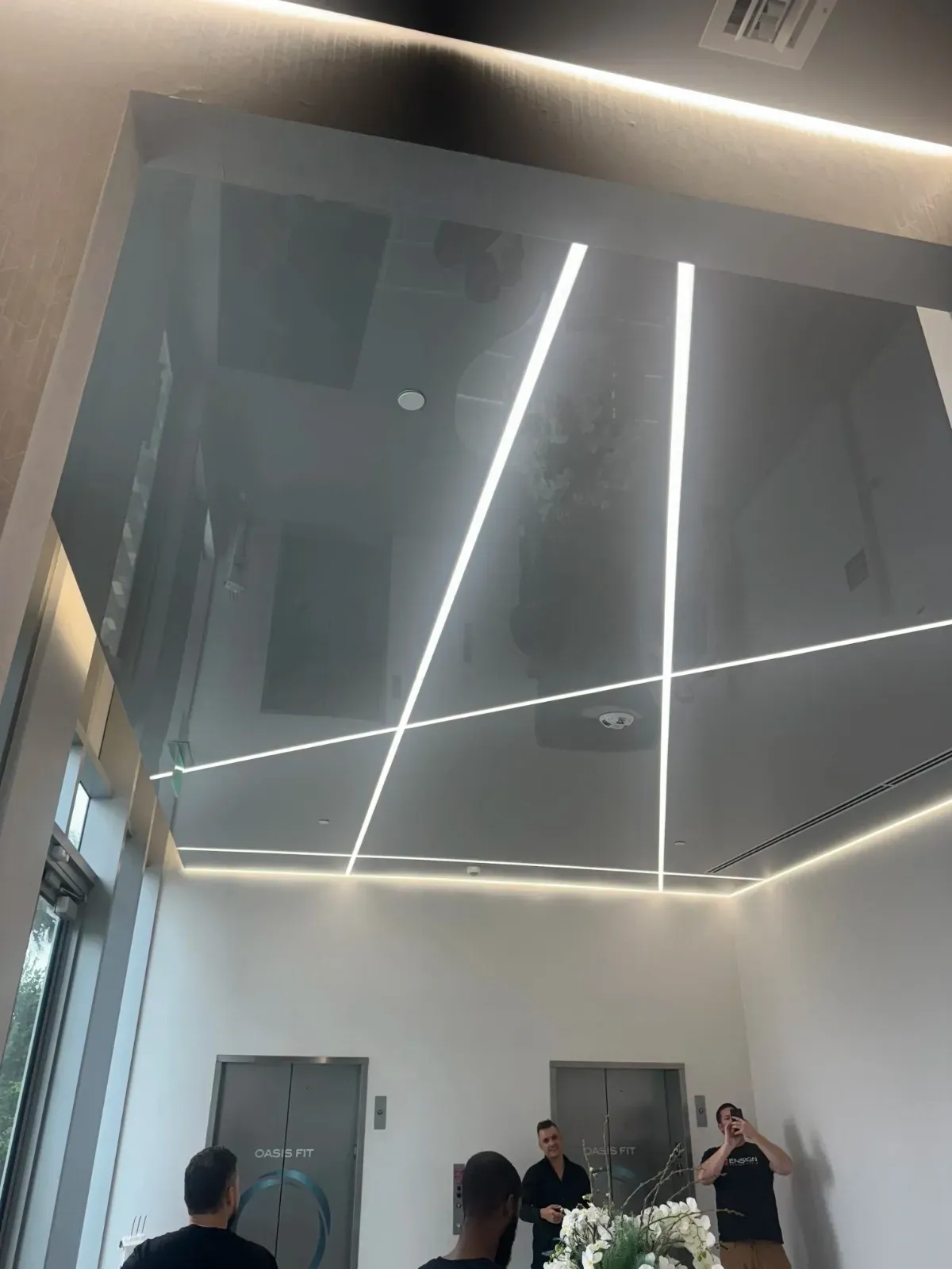 A high, glossy, reflective black stretch ceiling installation in a modern, well-lit indoor space (possibly a lobby or entrance), featuring two bright diagonal linear lights crossing the center and a linear perimeter light. Reflections of people and text on the wall below (like 'OASIS FIT') are visible in the ceiling.