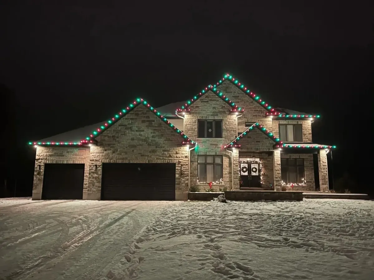 A large, two-story stone house at night, decorated with alternating red and green LED lights outlining the roofline and gables, with snow covering the front yard and driveway.