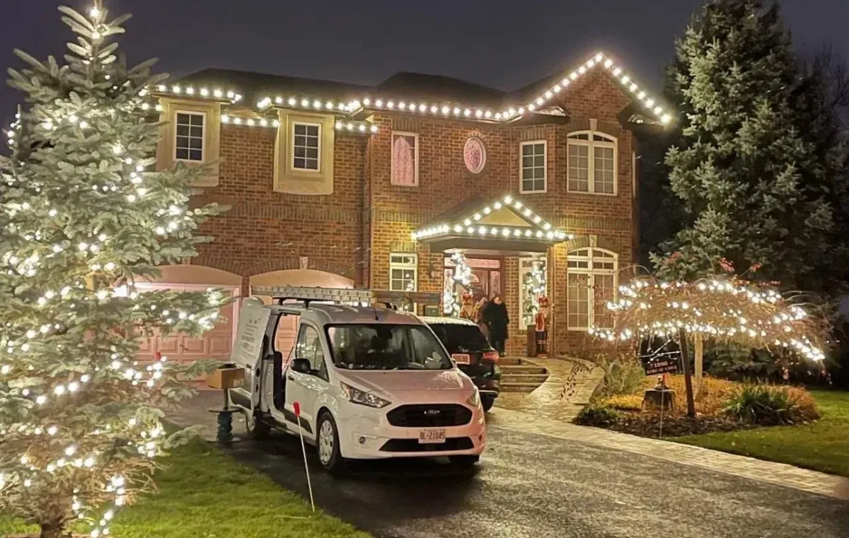 A luxurious brick home at night beautifully decorated for the holidays with warm white lights outlining the roofline, windows, and front porch. A white service van is parked in the driveway next to a large, decorated evergreen tree.