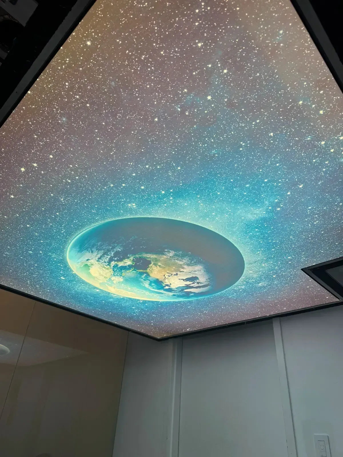 An interior ceiling featuring a stretch ceiling panel with a high-definition image of a vibrant, starlit outer space scene with a flat Earth-like planet or disk centered in the middle.