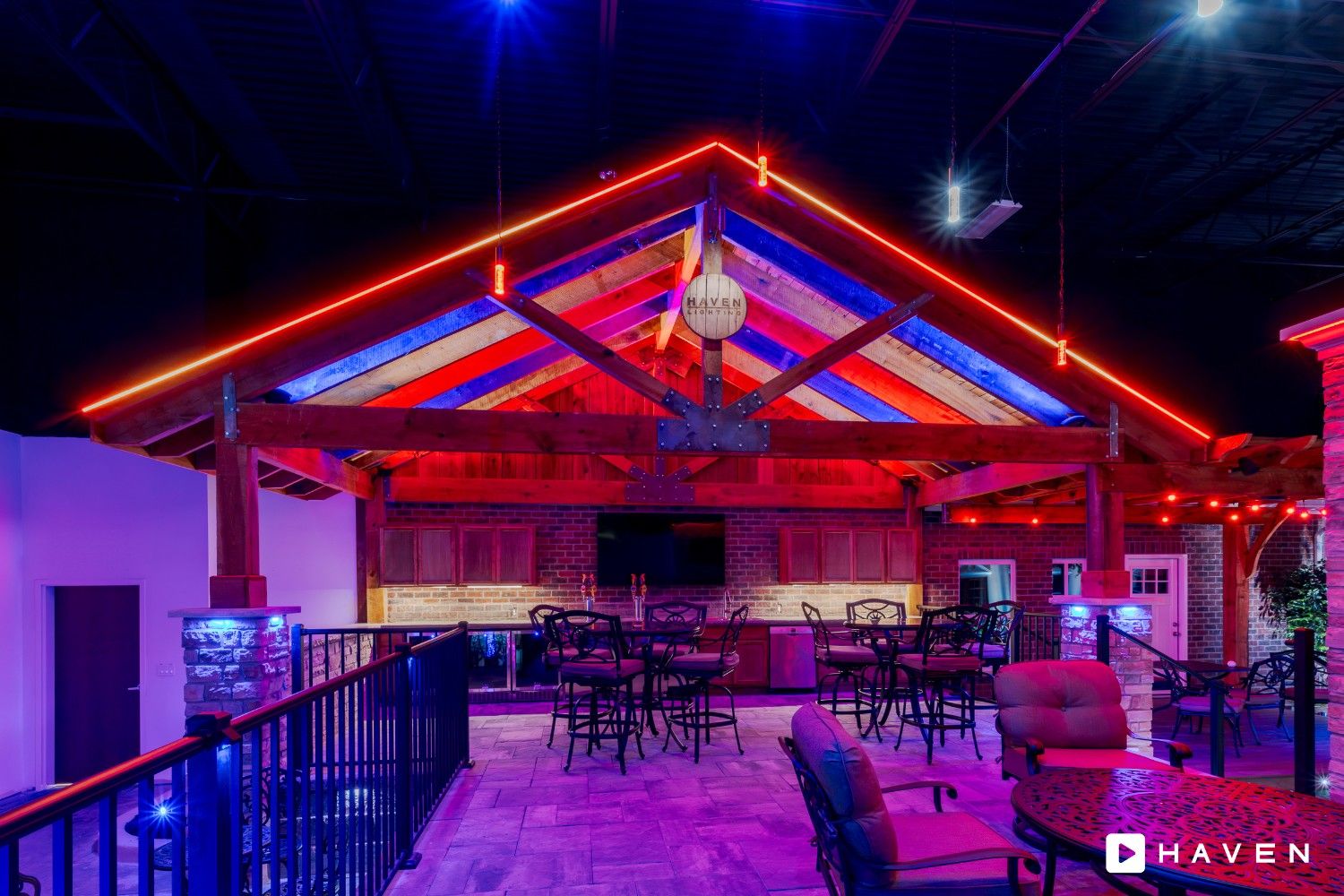 An interior/exterior covered bar and seating area with an exposed wooden truss ceiling brightly lit with contrasting red and blue colored lighting accents across the structure and seating area.