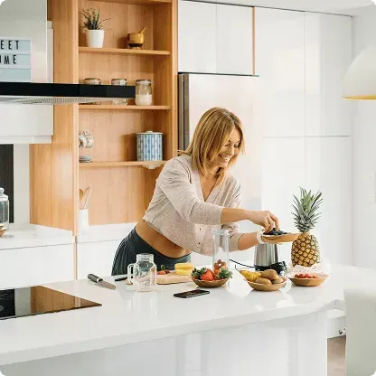 Smiling woman preparing a fruit smoothie in a modern white kitchen with fresh fruits on the counter.