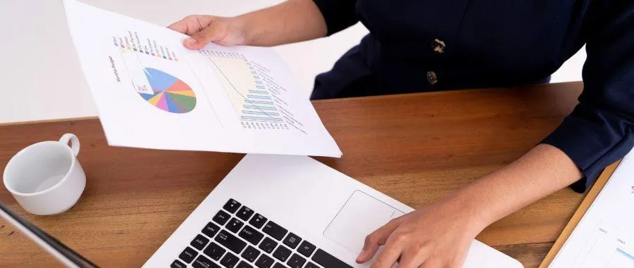 A desktop with mug and one hand on a laptop and the other holding a financial document.