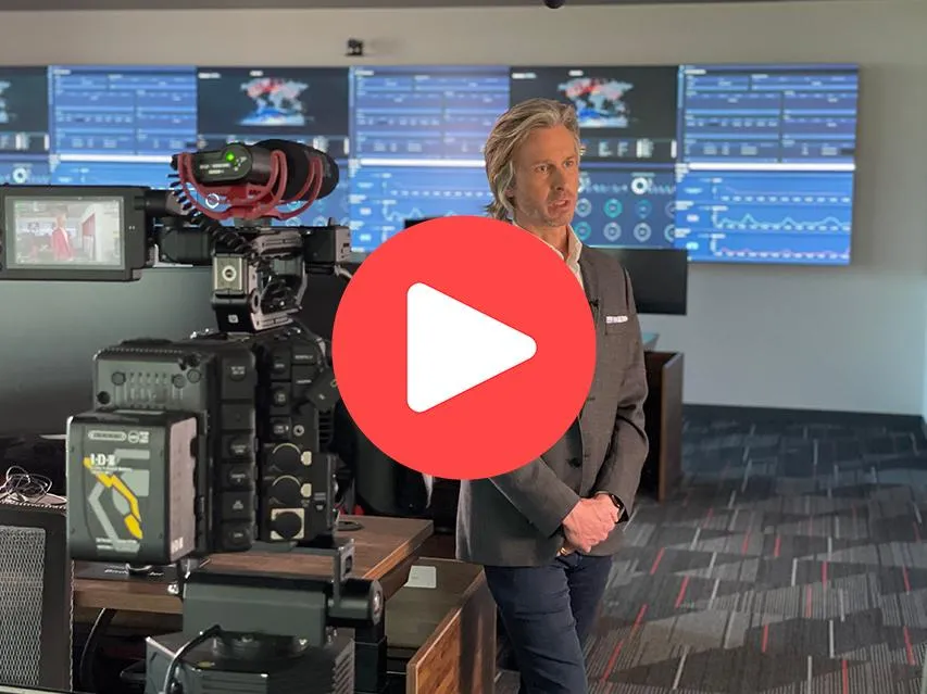 Man being interviewed for san antonio video shoot at bitedefender. He is standing in front of a wall of monitor and a large corporate video camera is pointed at him. There is a red play button centered on the image