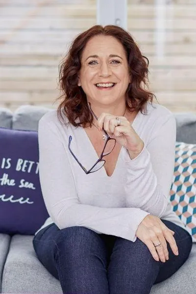 Photo of Emma Davis sat on sofa holding her glasses in one hand and smiling at the camera