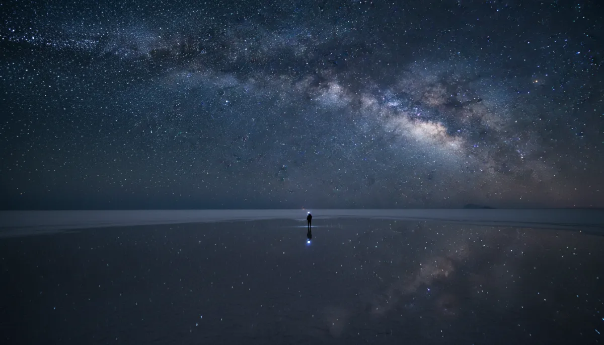 escena fotorrealista de larga exposición nocturna en el Salar de Uyuni, superficie blanca ligeramente inundada reflejando un cielo completamente estrellado, Vía Láctea nítida, silueta pequeña de viajero con linterna frontal, tonos fríos y neutros