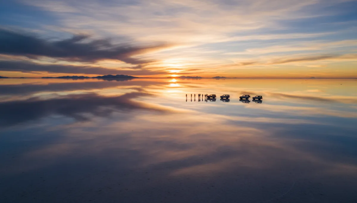 Vista aérea fotorrealista y cinematográfica del Salar de Uyuni en temporada de lluvias, gigantesco espejo de agua reflejando nubes dramáticas y luz dorada del atardecer, pequeñas siluetas de viajeros y 4x4 en la distancia, estilo National Geographic vibrante y emocional