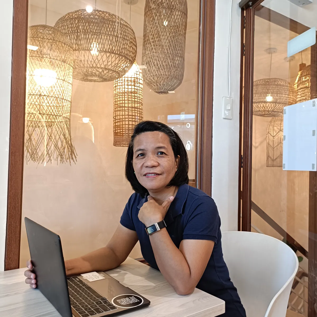 Divine Balubar seated at a table with an open laptop and warm woven pendant lights in the background