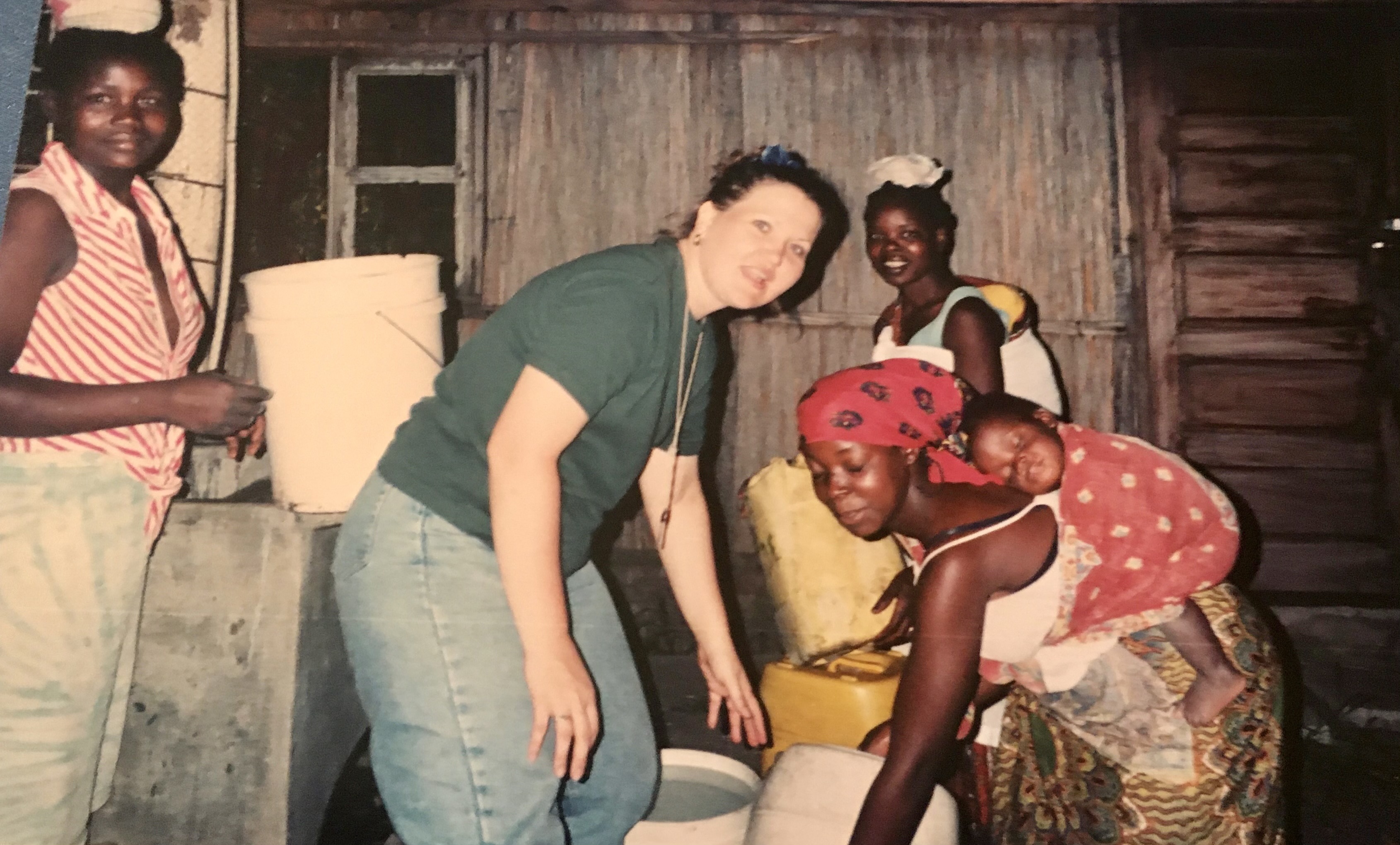 A cheerful interaction between a white woman and an African woman working together near a rustic wooden structure and water buckets.