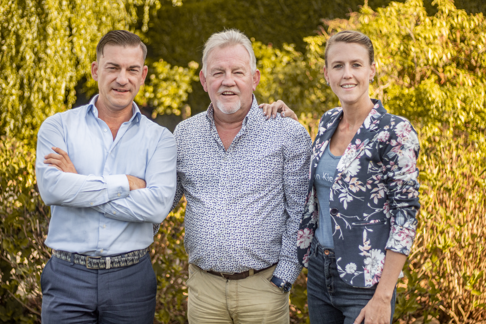 Driepersoons familieportret van het team van het makelaarskantoor buiten het kantoor, warme natuurlijke lichtinval, vriendelijke gezichten, fotorealistische compositie die continuïteit en vertrouwen in lokaal financieel advies benadrukt.