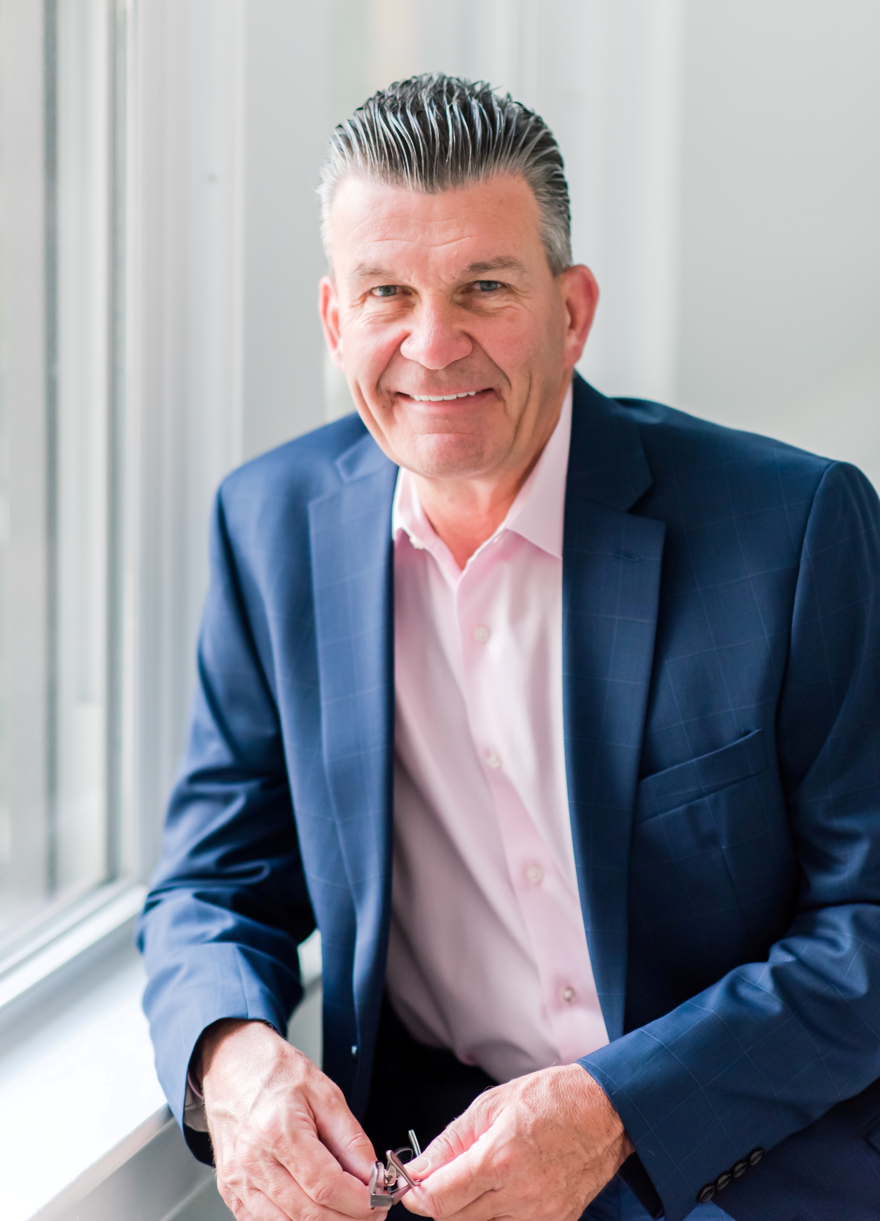 Professional portrait of John, smiling confidently in a modern office setting, wearing a navy suit and light blue shirt, with soft natural lighting and a blurred background of bookshelves and plants. 3:2 aspect ratio.