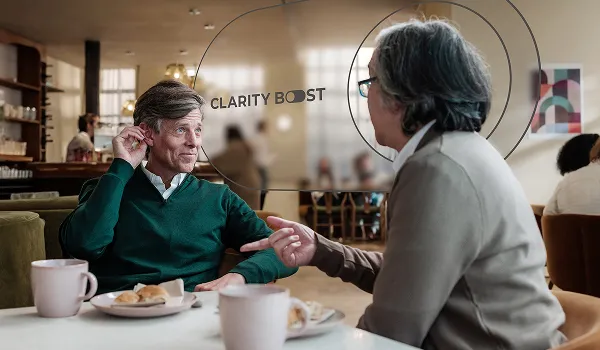 Over the shoulder view of a woman talking to a man in a bustling cafe, focusing on his hearing aid.