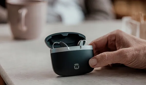 Sleek, dark grey pill-shaped charging case slightly open, revealing two hearing aids inside.
