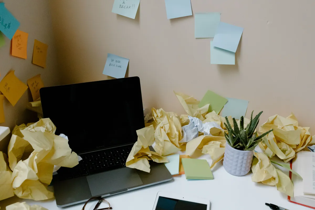 A messy desk setup with a laptop, notebooks, coffee mug, and many crumpled yellow paper balls. The wall behind the desk is covered in colorful sticky notes with questions like 'Module ideas?', 'Course structure?', 'What to include?', 'Next steps?', and 'Audience?'.