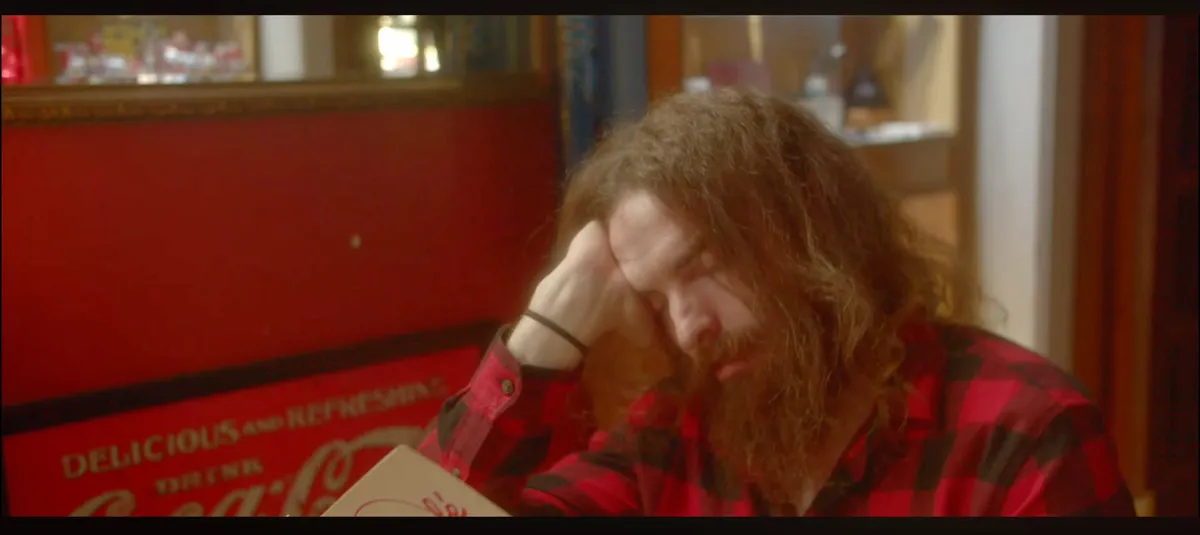 Film still from 3am Cafe — man in a warm-lit late-night café with vintage Coca‑Cola sign in foreground