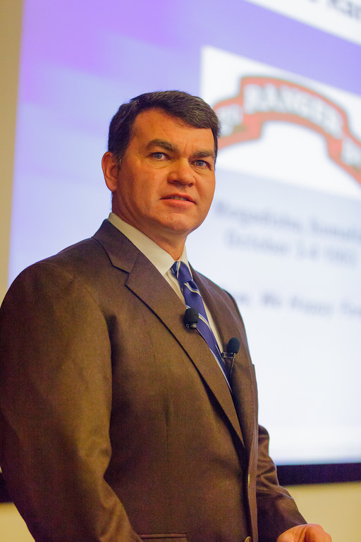 photo of James Lechner speaking at a podium, wearing a suit and tie, against a blurred background with a Ranger logo
