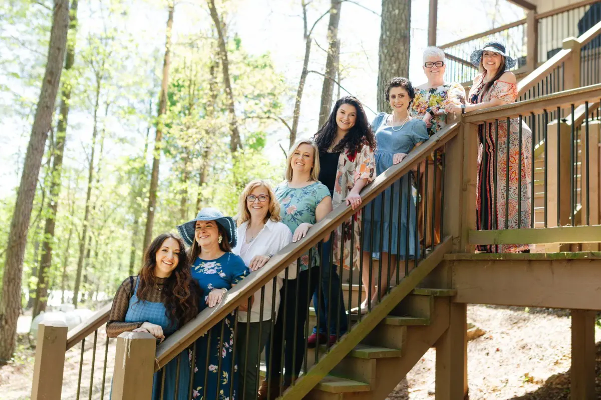 Five smiling women outdoors against a backdrop of trees, holding cameras, dressed in bright, colorful casual wear.