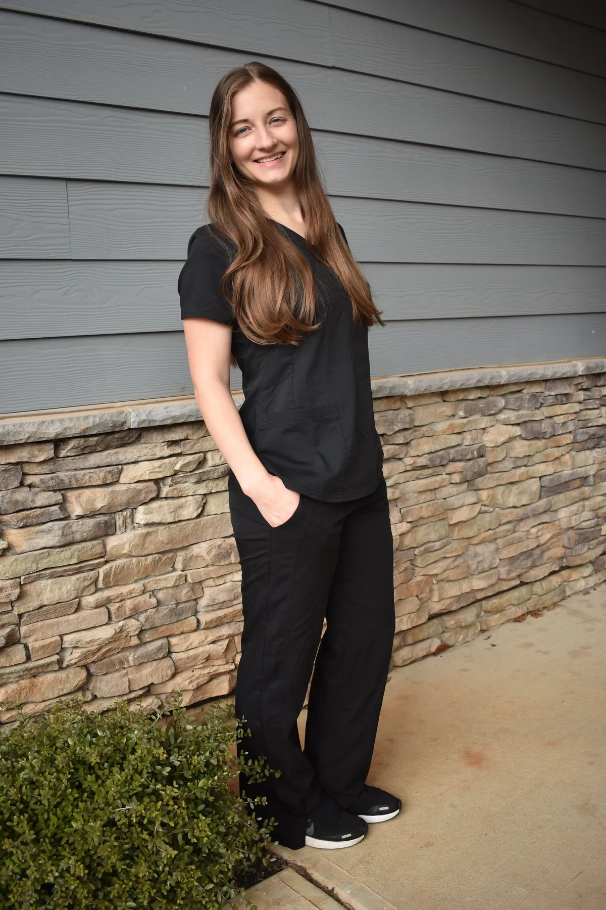 Coach standing outdoors against blue wall and stone siding, smiling with hands in pockets.