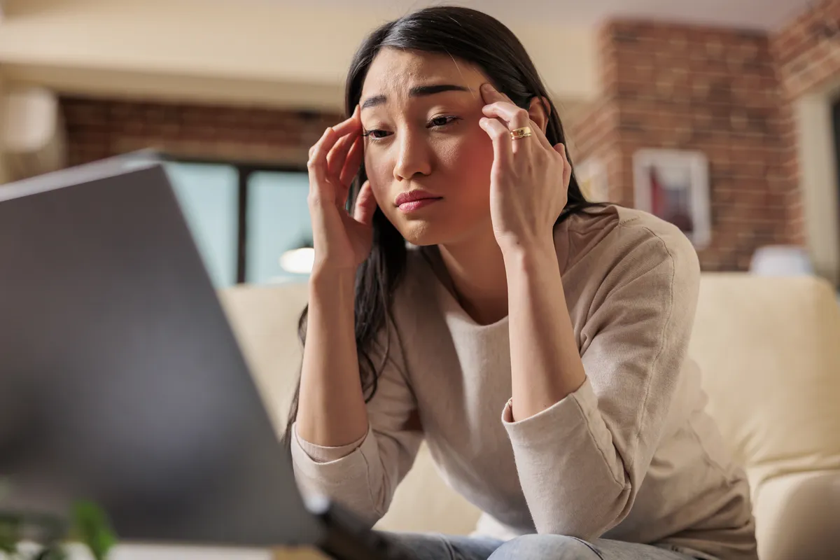 An Asian woman sitting at a laptop, looking frustrated and confused while trying to figure out what digital product to create