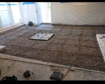 Looking down into an excavated dirt trench showing a newly poured square concrete footing pad next to an older foundation wall