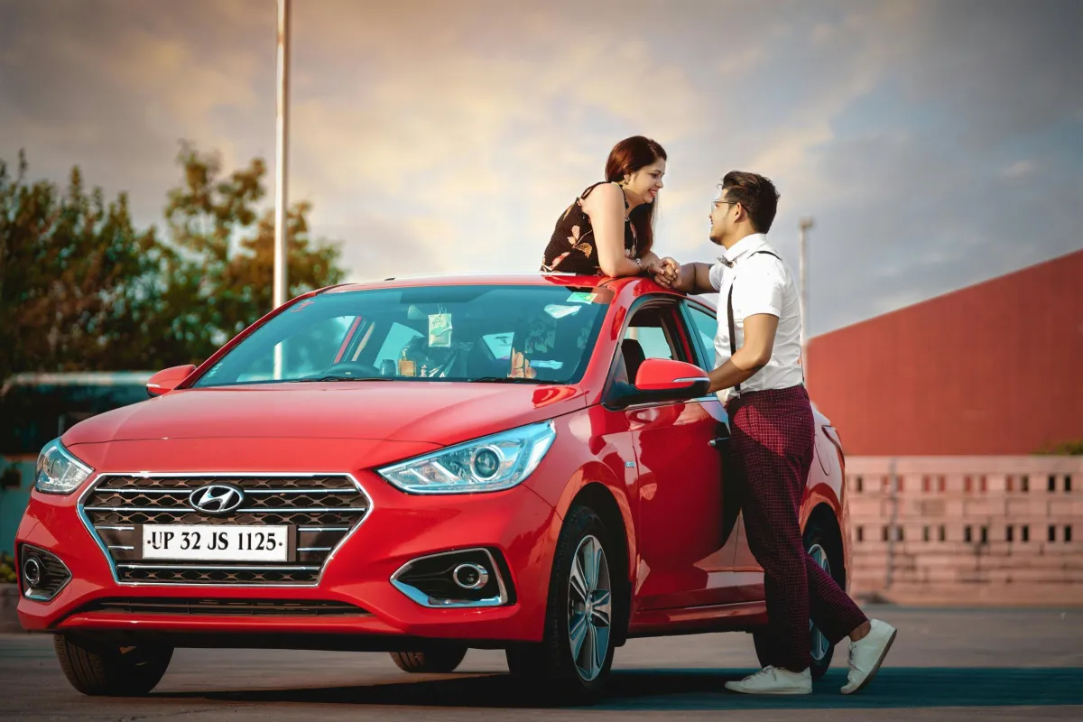 A man and a woman with love expressions, with the woman sitting on the roof of a red Hyundai car, and the man stands leaning on the car door, engaging in a romantic interaction in an outdoor parking lot during sunset.