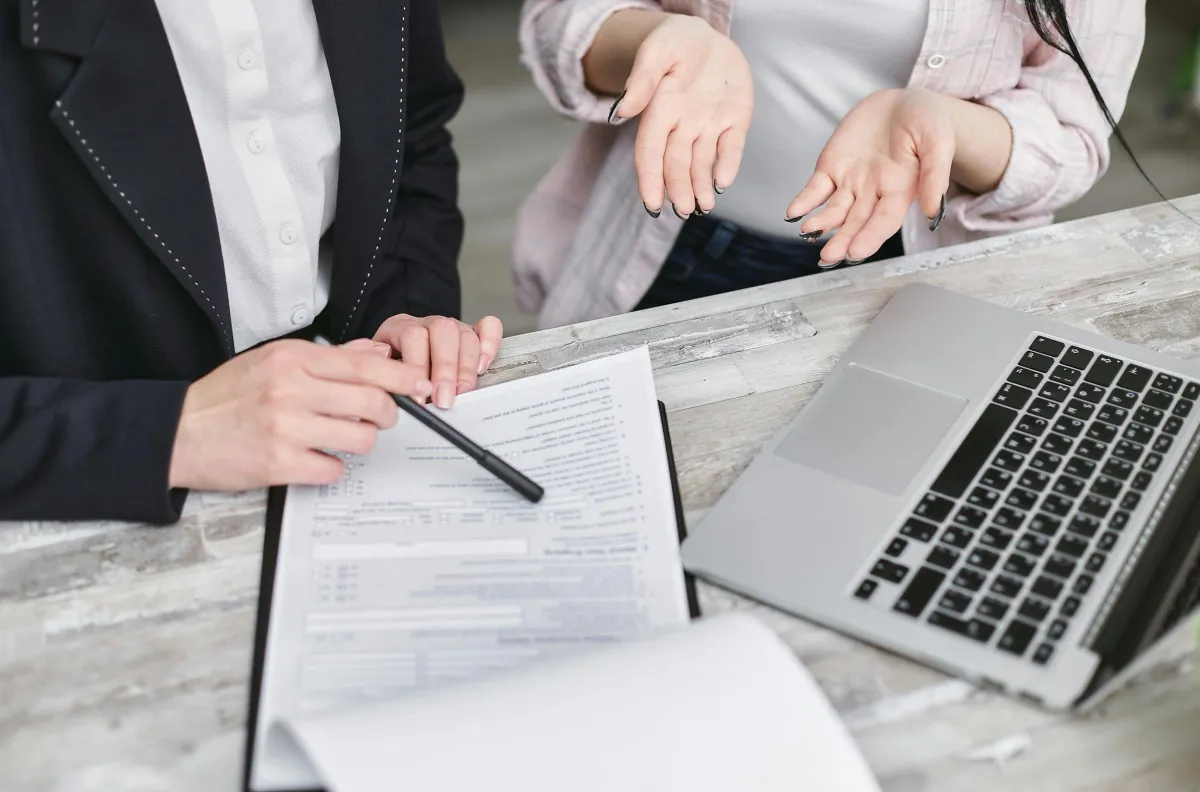 Two people discussing documents and a laptop on a wooden table.
