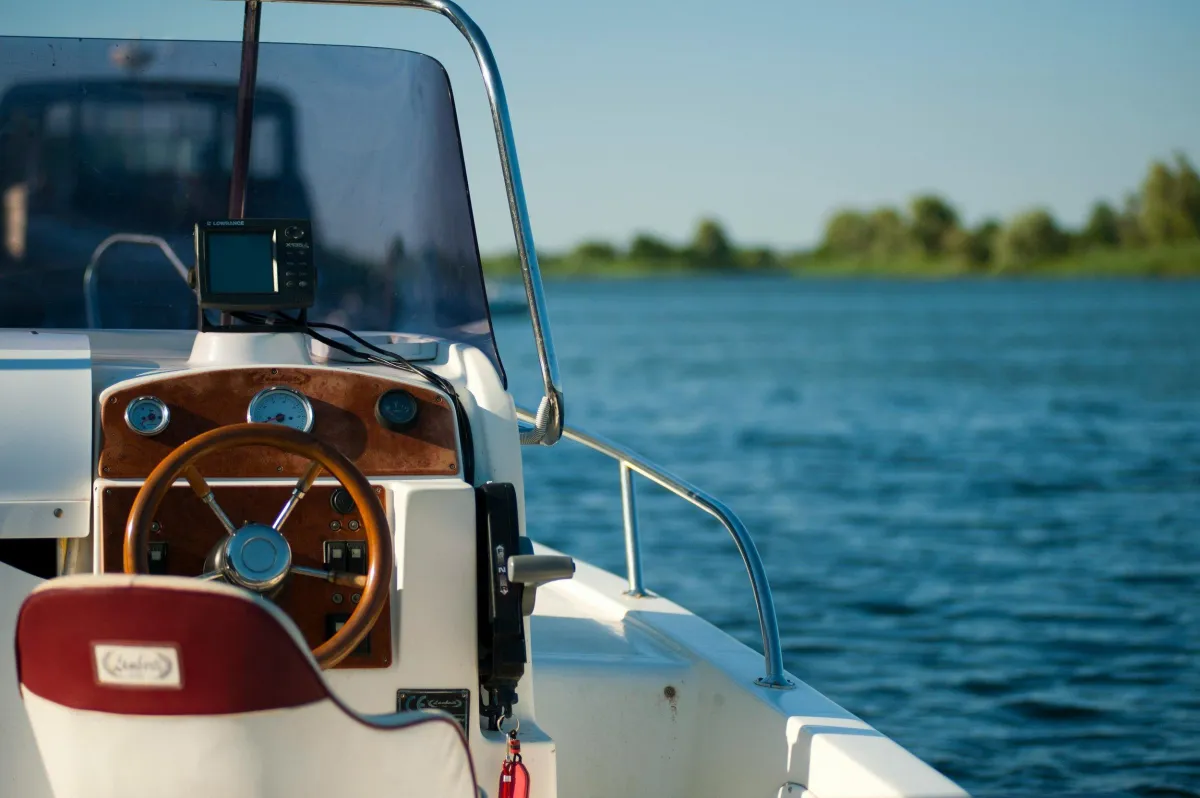 View of a boat's steering console with a woodgrain dashboard, gauges, and a steering wheel, on a body of water with a distant shoreline and trees in the background.