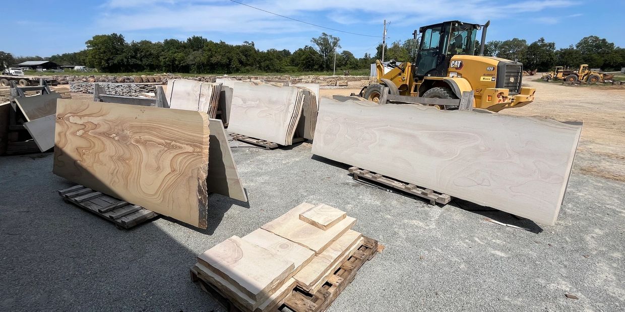 Large slabs of stone are displayed outdoors on pallets with construction equipment nearby.