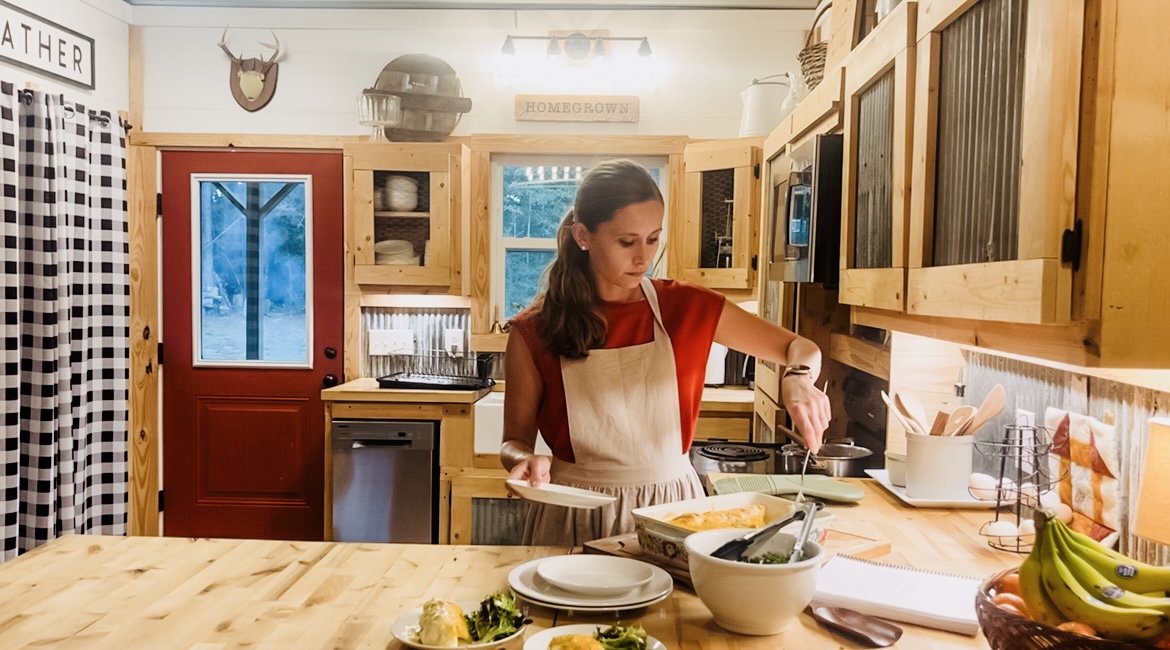 A woman in a red shirt and beige apron cooking in a rustic wooden kitchen, with a bowl of salad, plates, and bananas on the counter.