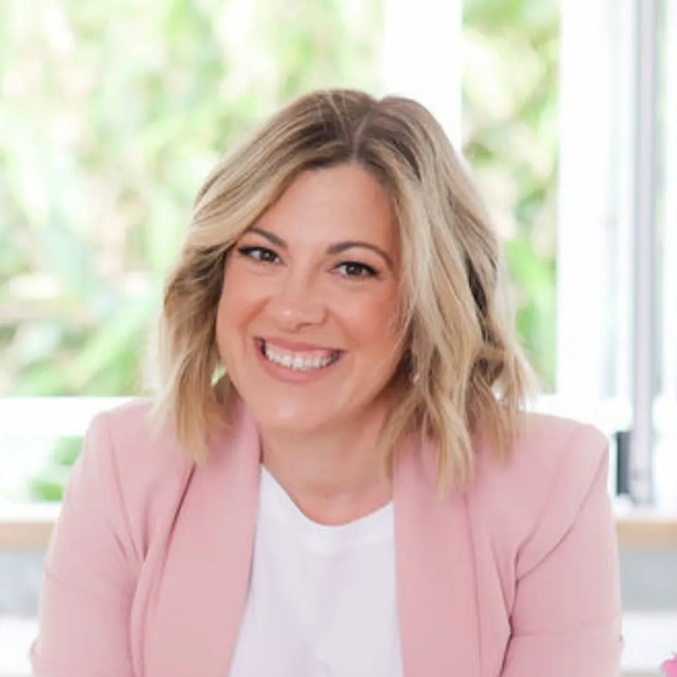 Smiling blonde woman wearing a pink top, sitting at a white desk and looking at an iPad, with soft floral elements in the background