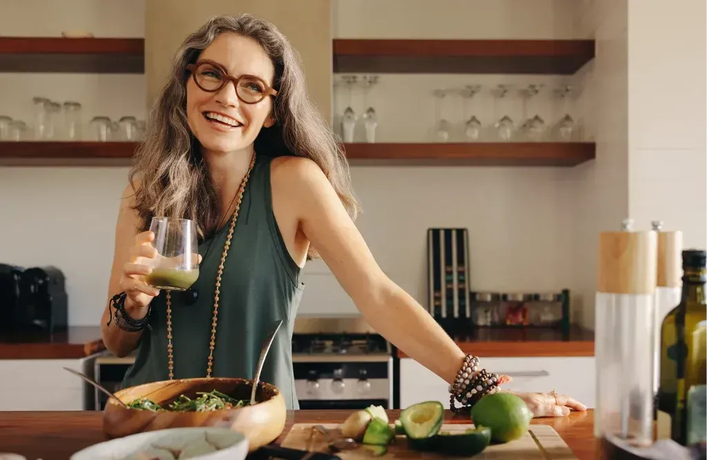 Woman smiling in the kitchen holding a glass of wine with vegetables on the counter