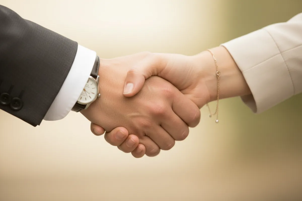 Two hands—one male, one female—shake in a close-up, symbolizing trust. The background is softly blurred in beige and cream, with subtle green accents. The image conveys partnership, reliability, and mutual respect in a professional context.