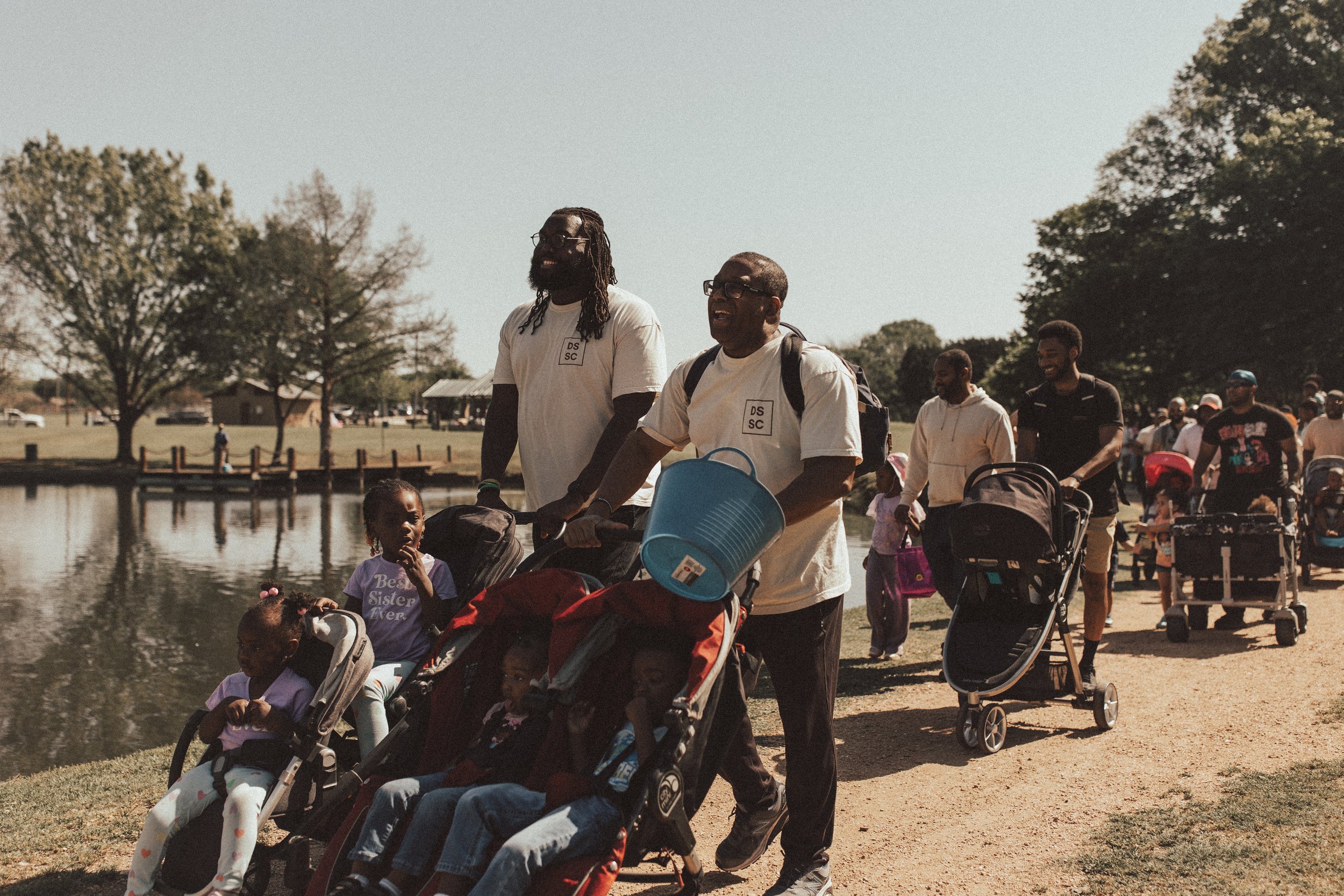 People walking along a path near a lake, pushing strollers and surrounded by trees on a sunny day.