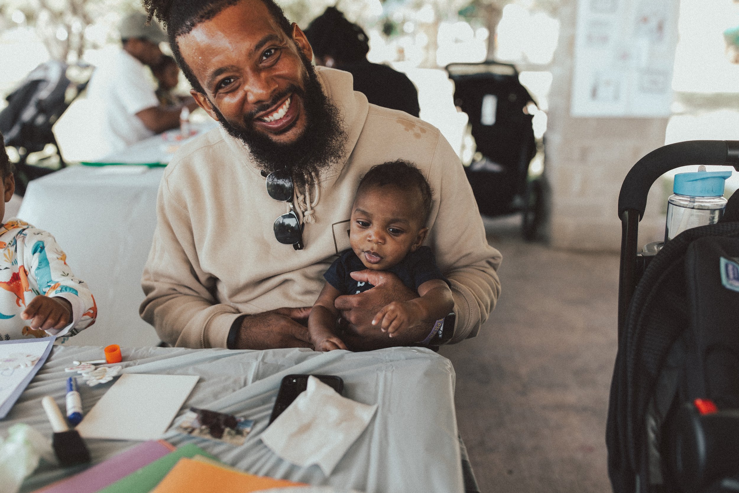 A smiling man with a beard and sunglasses hanging from his shirt collar holding a baby with short hair, sitting at a table with art supplies, in a bright indoor space with other people in the background.