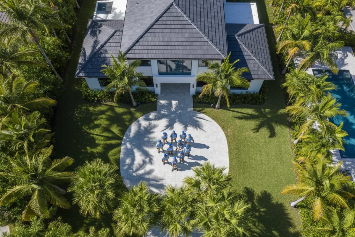 Aerial view of a modern South Florida home with a new dark tile roof, palm trees, and a team of diverse construction professionals in branded uniforms, bright sunlight, clean lines, contemporary style, 3:2 aspect