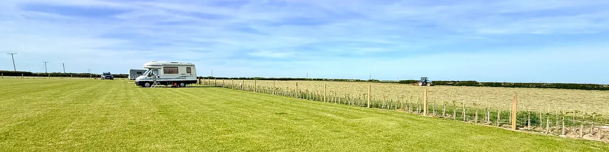 A wide grassy touring field at Bungalow Farm in Buckton with a motorhome parked under a bright blue sky, overlooking open farmland and a tractor working in the distance.