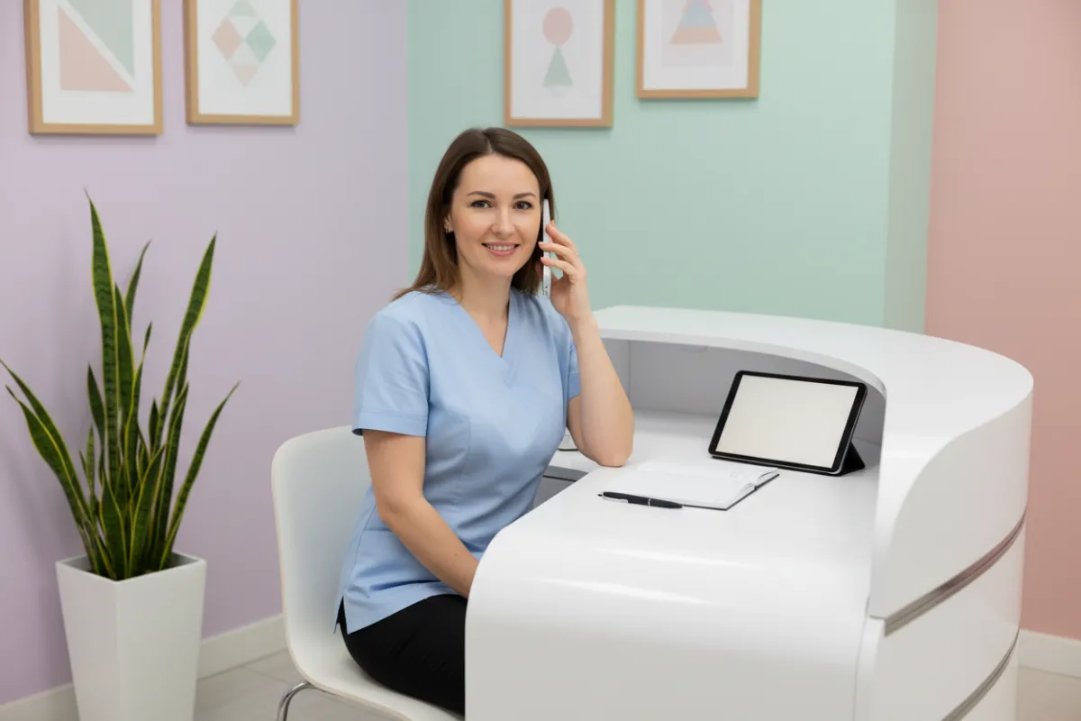 Clinic staff member answering a phone call at a modern desk, with a digital device nearby and a friendly expression, in a softly lit office.