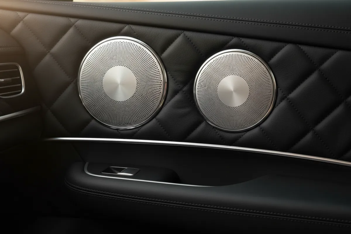 Close-up of a car door panel featuring flush-mounted premium speakers with brushed aluminum grilles, set against a black leather interior. The lighting emphasizes the texture and quality of the materials used.