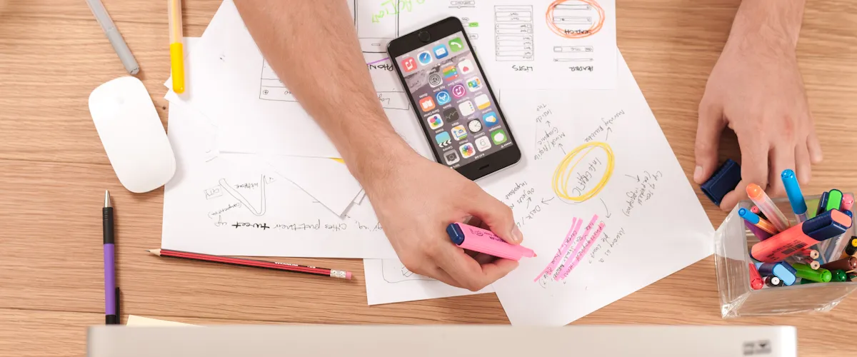 Hands using a pink highlighter on marketing strategy notes, smartphone displaying social media apps, and colorful pens in a workspace, emphasizing social media advertising for healthcare practices.