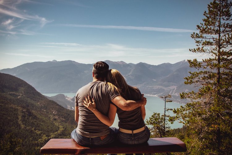 A couple sitting on a wooden bench, embracing each other while overlooking a mountainous landscape with a lake and trees.