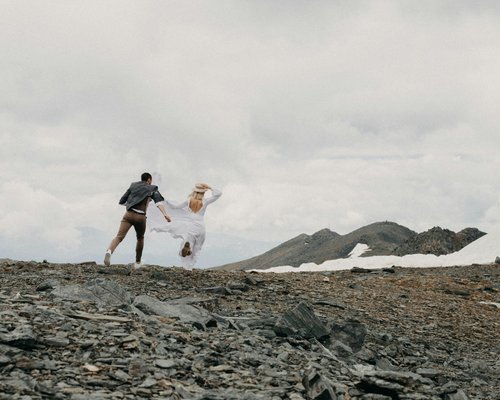 A man and woman running across a rocky, mountainous terrain under a cloudy sky, with snow patches visible on distant peaks.