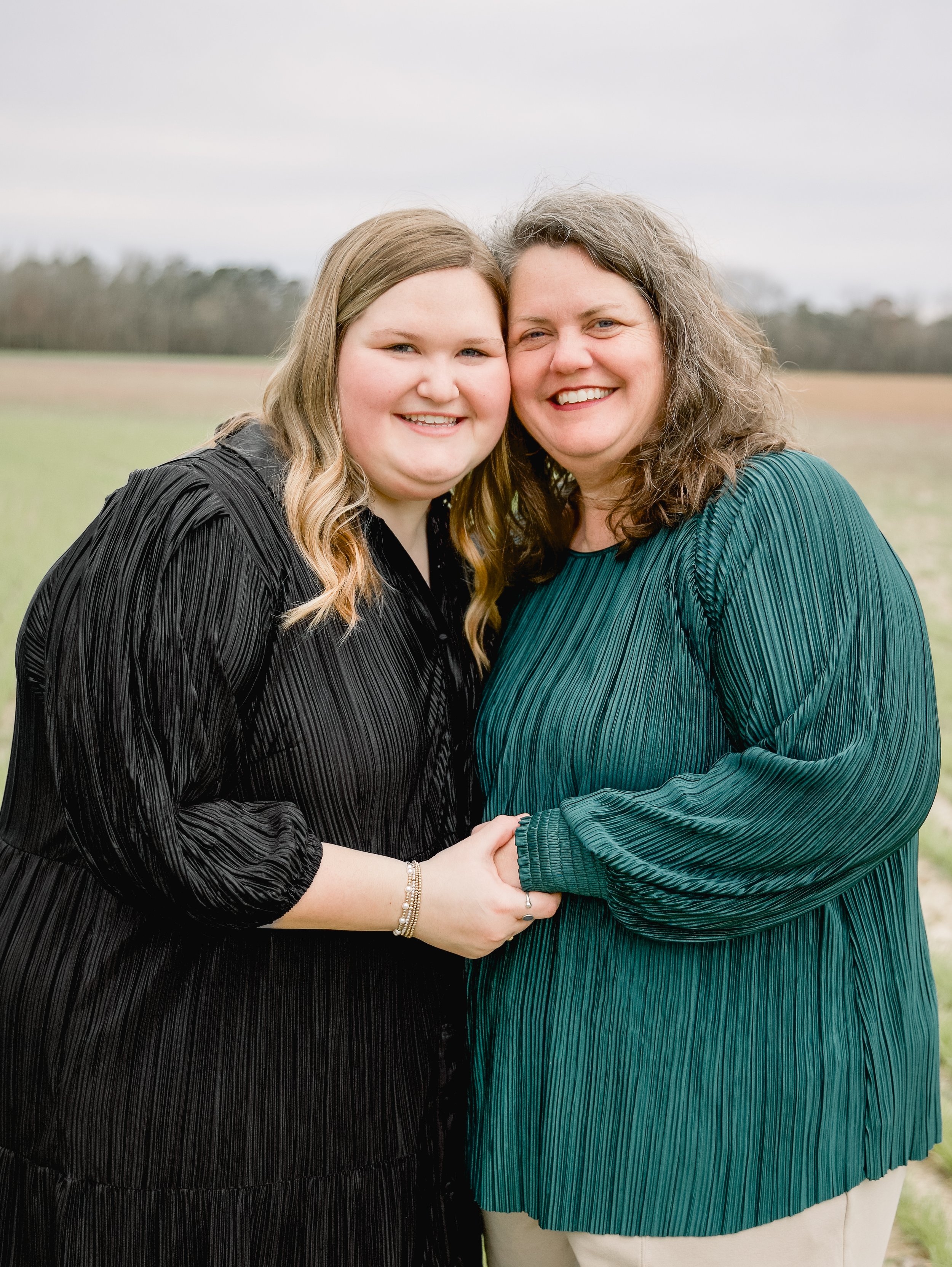 Two women standing outdoors, holding hands, smiling, with a field and overcast sky in the background.